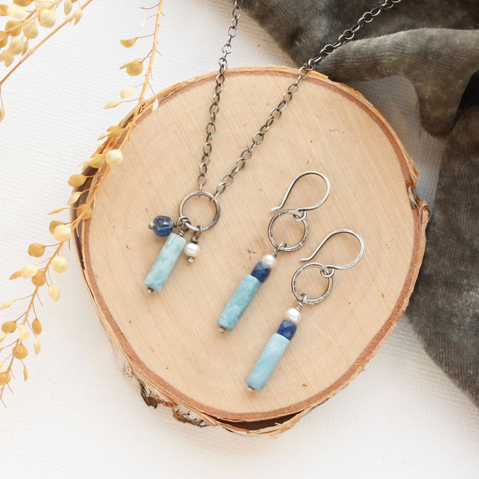 The mirage sydney charm necklace and matching earrings styled on a wooden round, gray velvet, and dried grass.