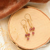 The garnet trio gold hoop earrings styled on wood with tan linen, brown velvet, and dried grass