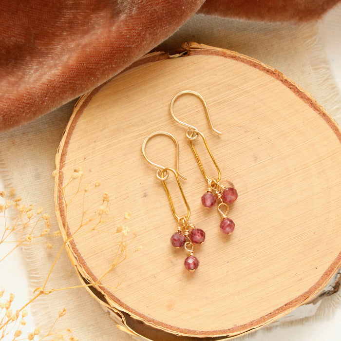 The garnet trio gold hoop earrings styled on wood with tan linen, brown velvet, and dried grass