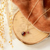 The garnet charm necklace styled on wood and tan linen with brown velvet and dried grass