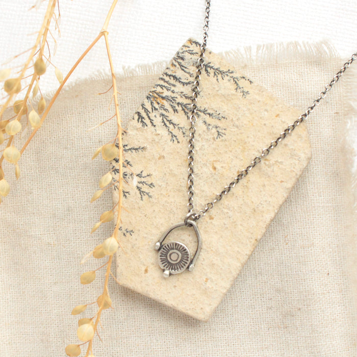 The mini talara shield necklace styled on a tan rock with linen and dried grass