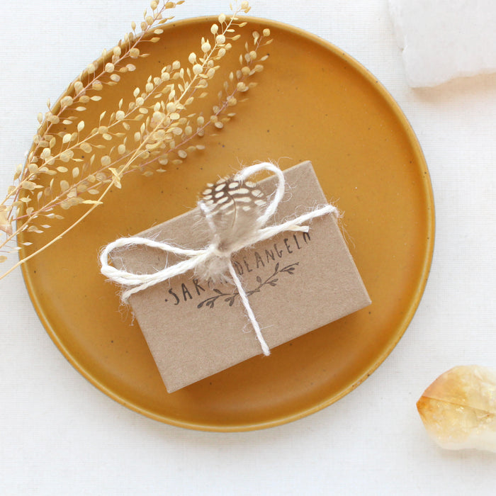 Sarah DeAngelo Jewelry box with signature gift wrap and feather styled on a yellow plate with dried grass and a crystal rock