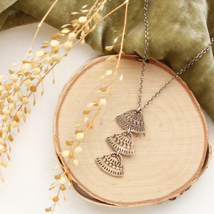 The bronze asmi trio triangle necklace styled on wood with green velvet and dried grass