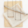 The bronze open bar necklace and silver open bar necklace styled together on a tan rock with tan linen and a wooden stick