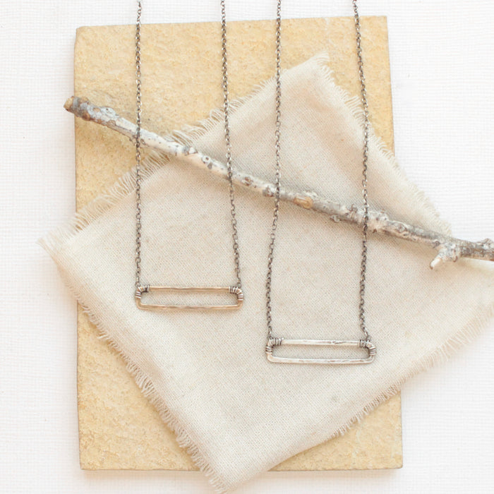 The bronze open bar necklace and silver open bar necklace styled together on a tan rock with tan linen and a wooden stick