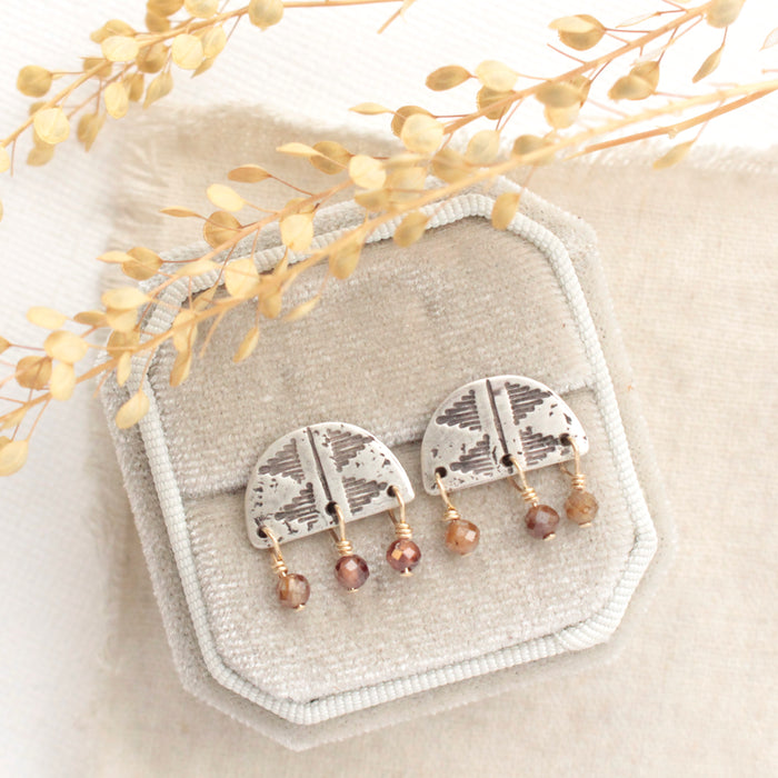 The brown zircon mixed metal pakal arch post earrings styled on tan linen with dried grass