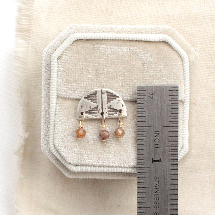A brown zircon pakal arch post earring next to a ruler for size reference