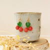 The little cherry earrings styled on a ceramic cup with wood and dried grass