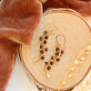 The garnet wrapped gold vine earrings styled on wood with orange velvet and dried grass