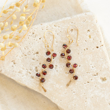 The garnet wrapped gold vine earrings styled on a white rock with linen and dried grass