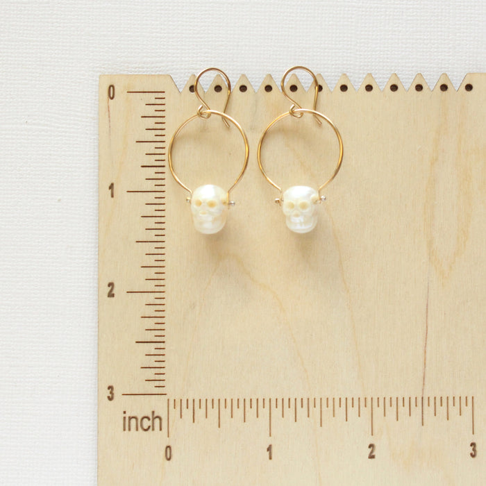 The carved pearl skull hoop earrings on a wooden ruler for size reference