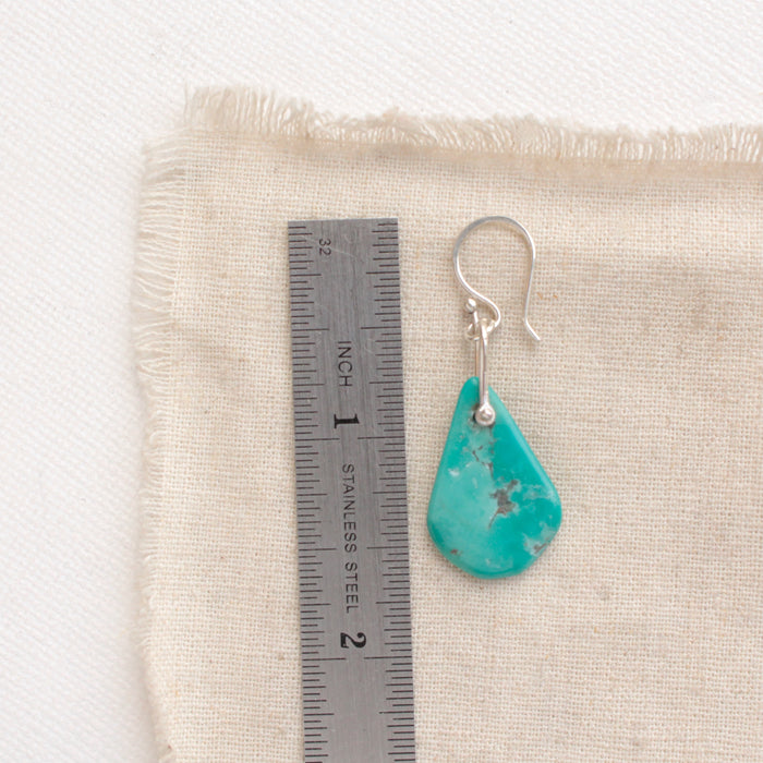 A pinned turquoise earring next to a ruler for size reference
