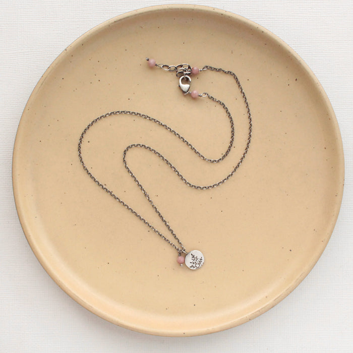 The rhodonite and sprout charm necklace styled on a tan plate to show the adjustable lobster clasp closure