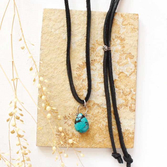 The turquoise drop adjustable leather necklace styled on a tan rock with dried grass