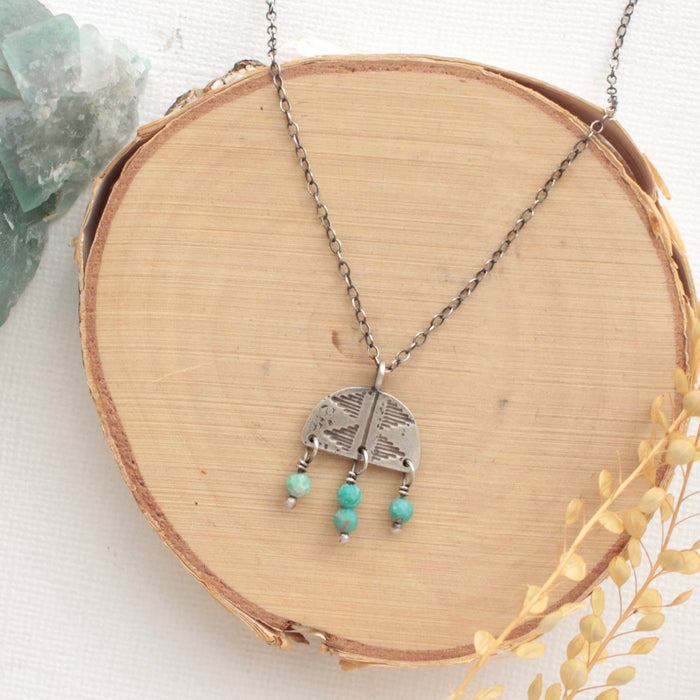 The turquoise fringe pakal arch necklace styled on wood with a green rock and dried grass
