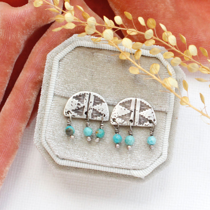 The turquoise pakal arch post earrings styled on a gray velvet box with orange velvet and dried grass