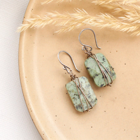 The wrapped african jasper earrings styled on a tan plate with dried grass