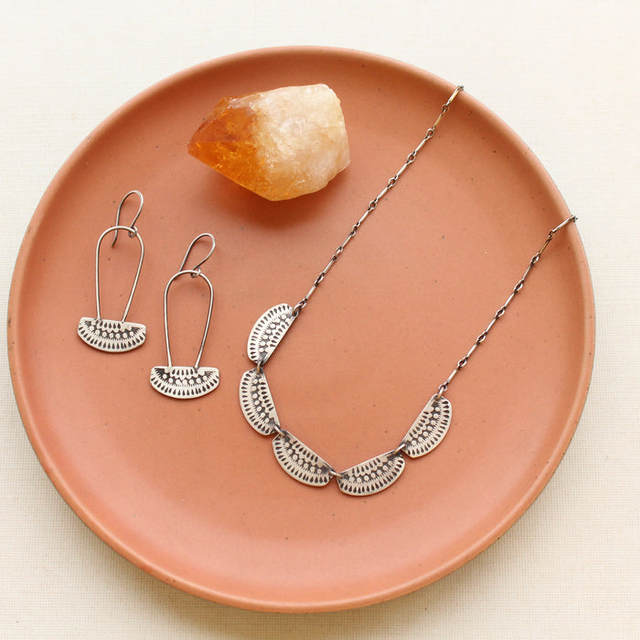 The asmi 5 collar necklace and asmi long loop earrings style on a red plate with a crystal