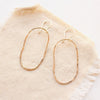 Gold organic shaped oval hoops with a hammered texture styled on tan linen.