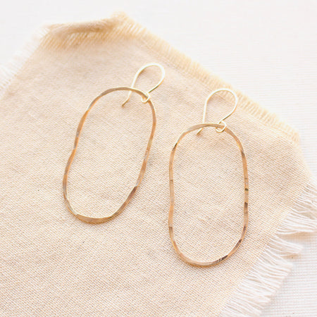 Gold organic shaped oval hoops with a hammered texture styled on tan linen.